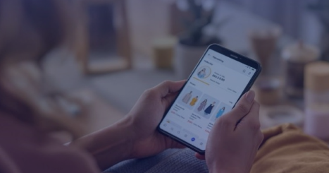 Person holding a smartphone while browsing an online shopping app displaying a profile page with several dresses for sale. The scene has a cozy indoor background with warm lighting and blurred decor elements