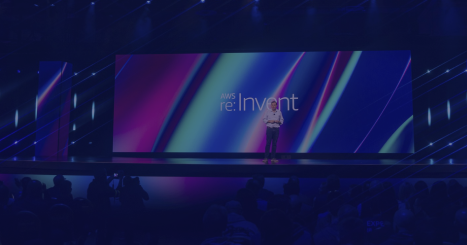 A presenter stands on stage in front of a large colorful screen displaying the AWS re:Invent logo during a keynote session.